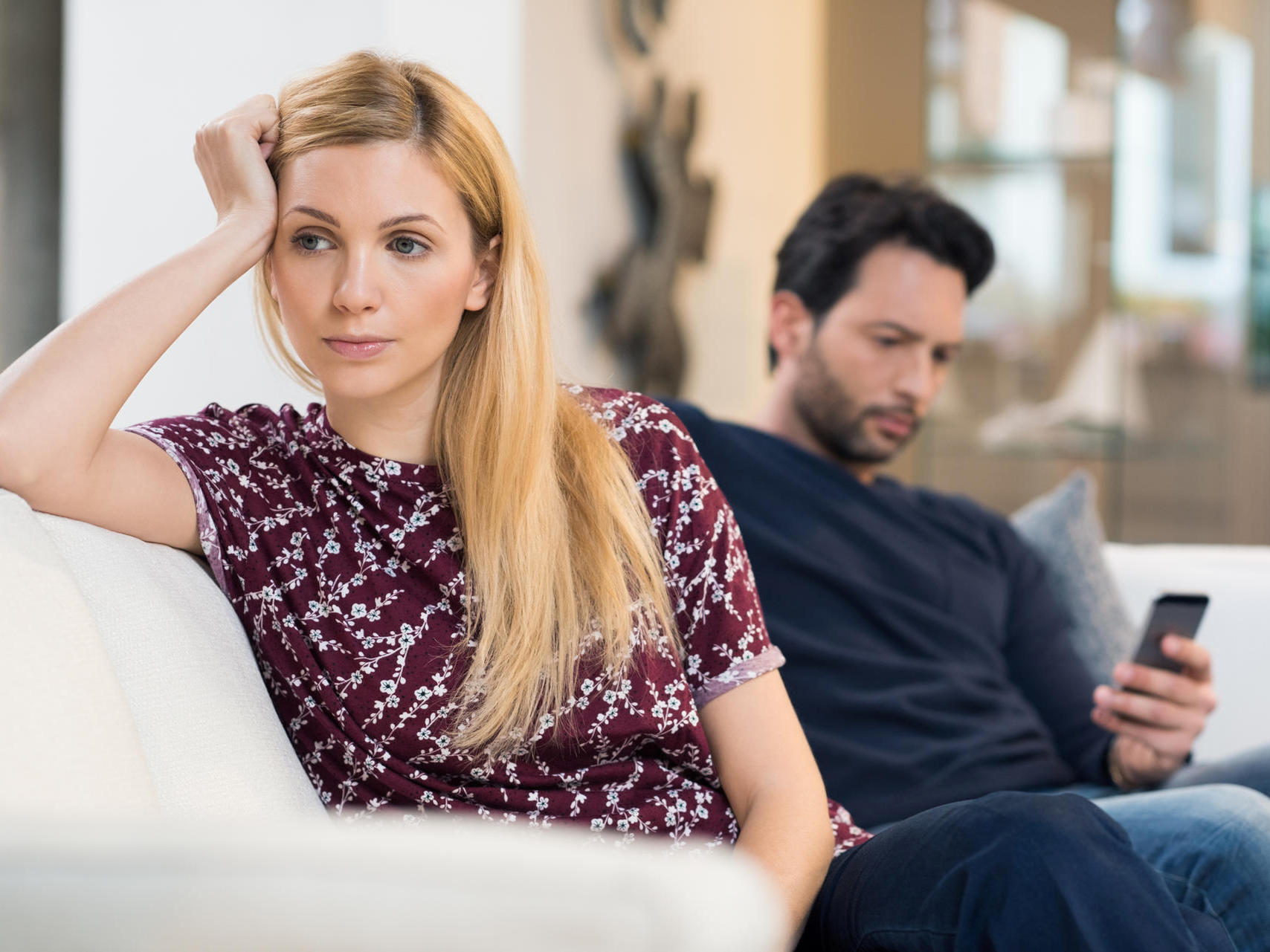 Young woman getting bored while man using phone in the background. Beautiful young woman feeling annoyed as man texting on phone. Young woman after an argument with her boyfriend in their living room.